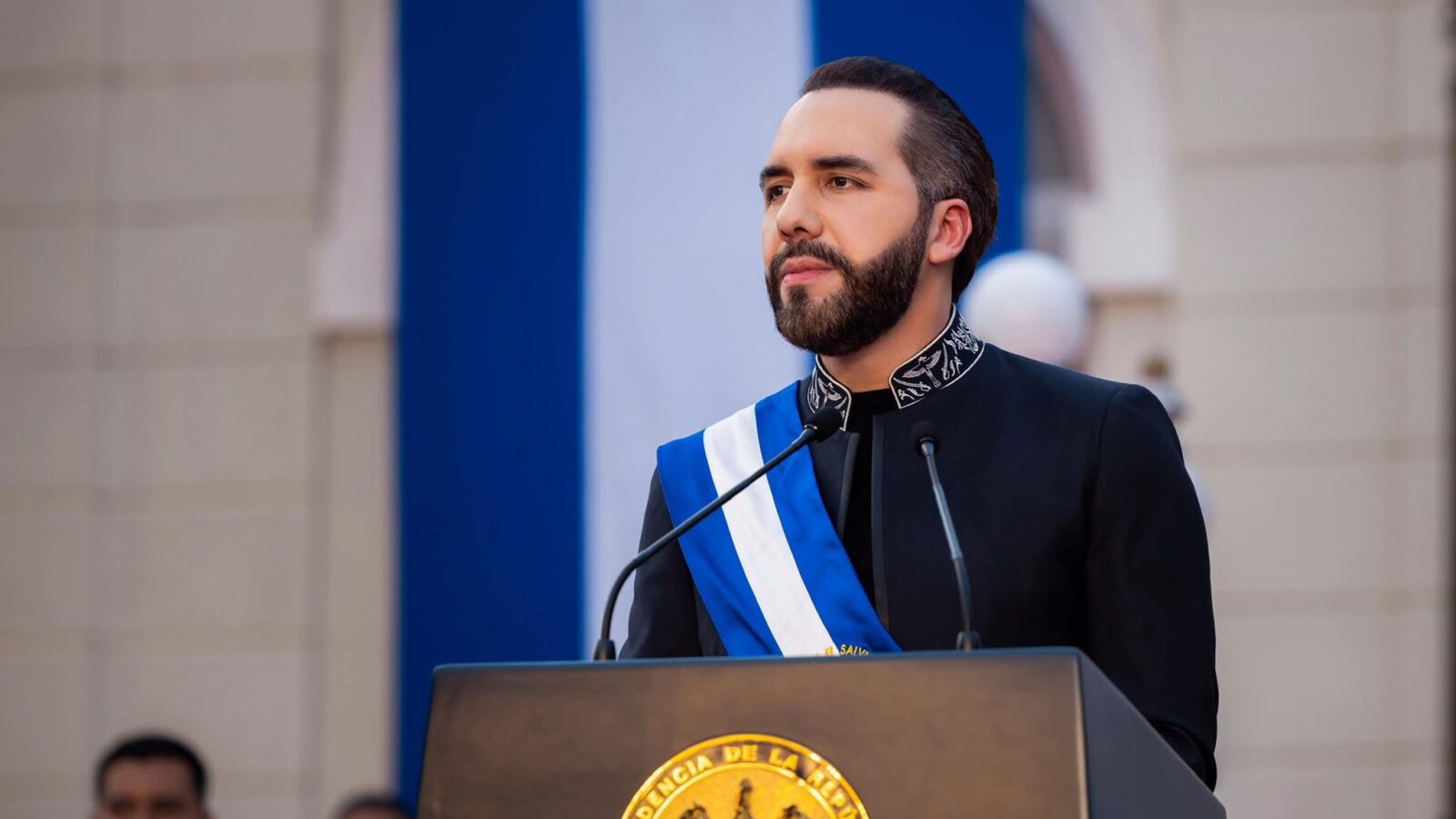Nayib Bukele durante un discurso, con la bandera salvadoreña de fondo, simbolizando su postura soberanista. Su rechazo al "wokismo" y políticas de género divide opiniones entre admiradores y críticos internacionales. (Foto: archivo X @nayibbukele)